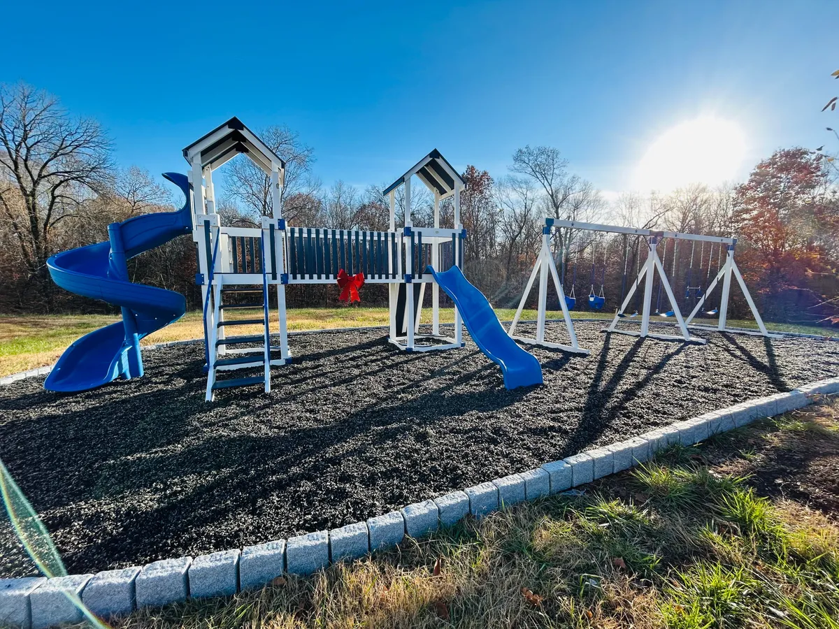 This image features an exterior scene centered on a playground structure with a blue slide and swings. The area is lively, likely situated in a residential community, and conveys a joyful, communal atmosphere with children playing and families gathering outdoors.