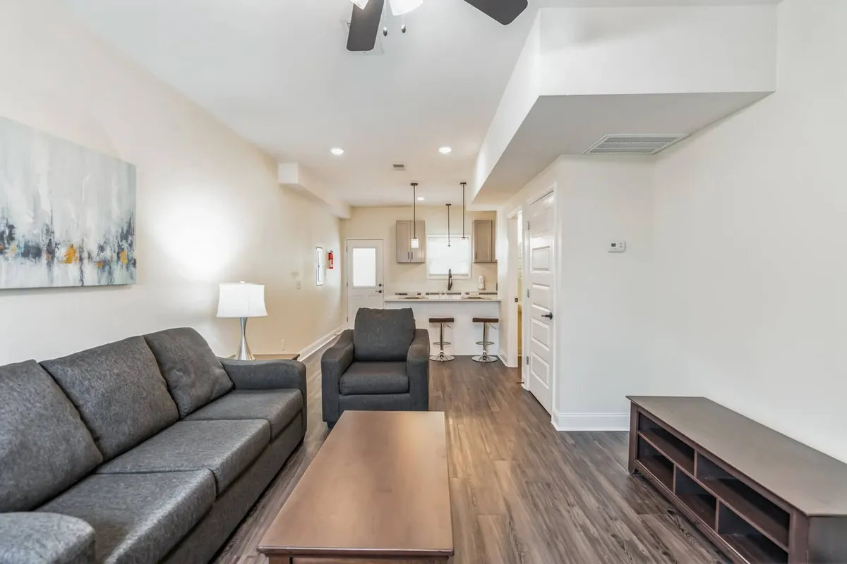 A cozy and welcoming living area that seamlessly connects to the kitchen, featuring a comfortable sofa, modern coffee table, soft lighting from a lamp, and a glimpse of the kitchen area. The decor strikes a balance between style and comfort, ideal for gatherings or quiet evenings.