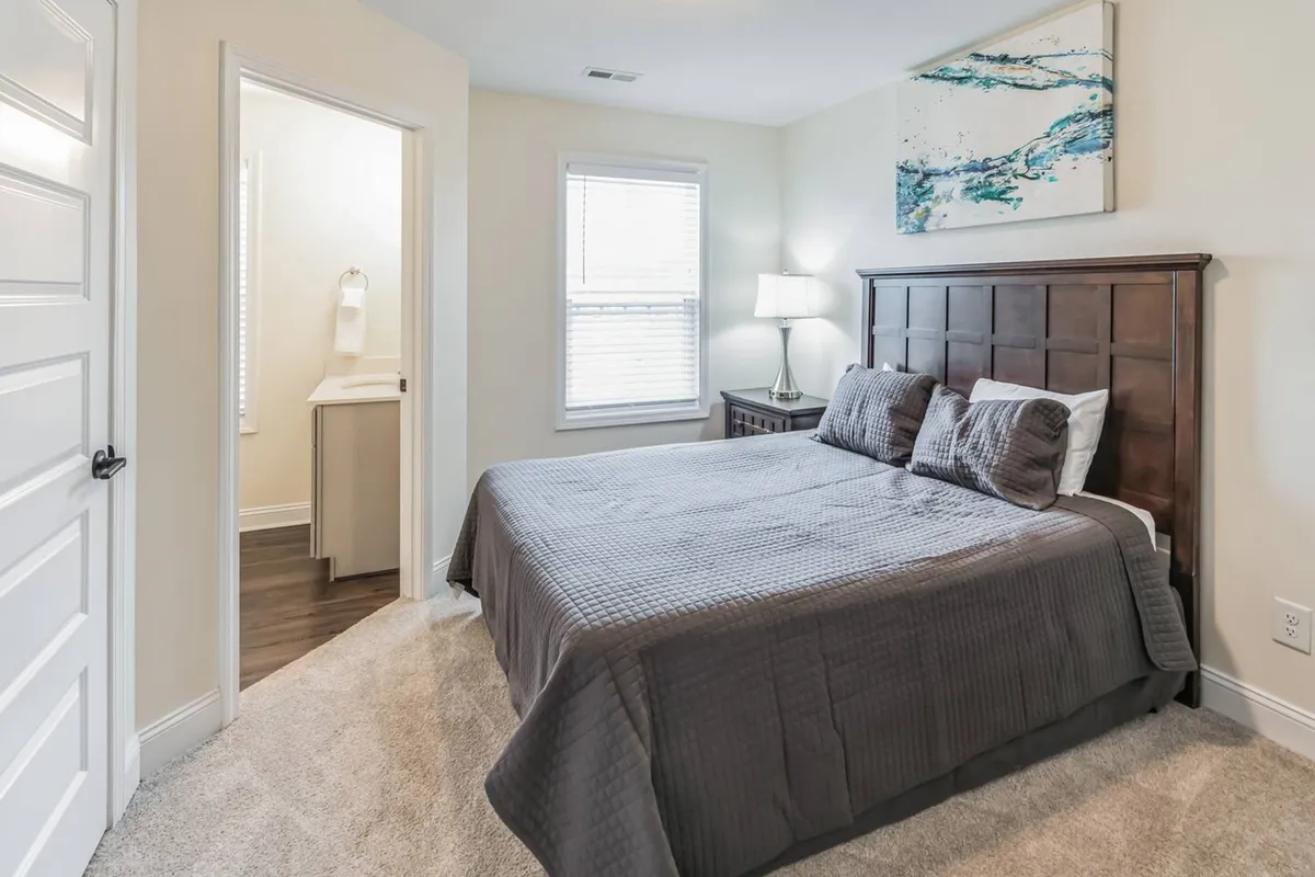 A tranquil bedroom with a bed draped in a gray quilt, complemented by a nightstand, lamp, and abstract wall art. The atmosphere is serene and restful, ideal for unwinding after a productive day.