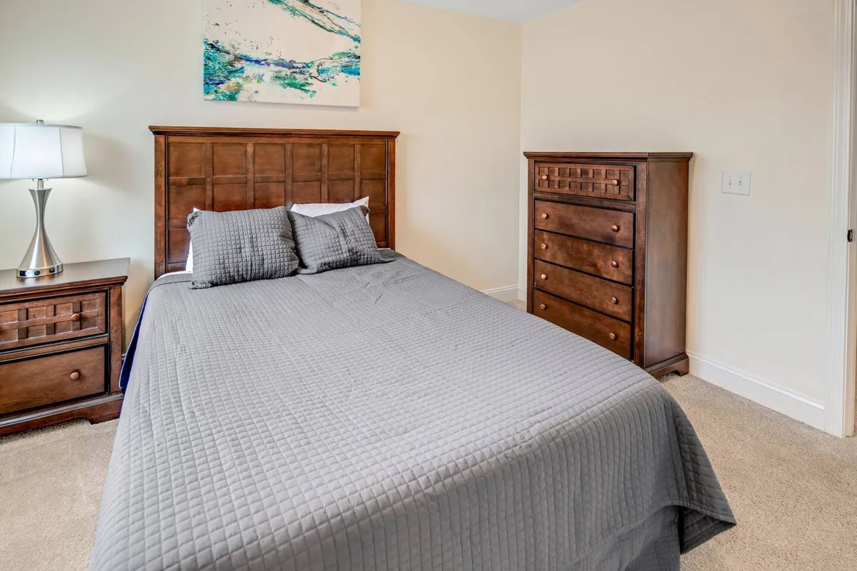 A serene bedroom with a gray-quilted bed, nightstand, lamp, chest of drawers, and an abstract painting. Sunlight streams softly into the space, emphasizing comfort and peace.