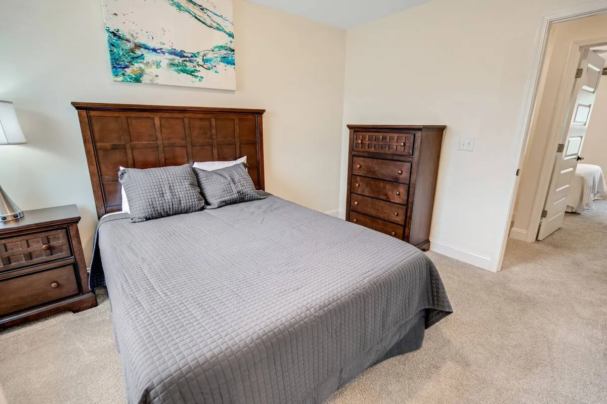 The image showcases a serene bedroom with a plush bed draped in a soft gray quilt, complemented by elegant wooden furniture and striking abstract wall art. Sunlight filters through the window, bathing the room in natural light and creating a tranquil, inviting atmosphere ideal for relaxation or reading.